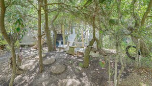 An outdoor seating area with Adirondack chairs, a wooden bench, and a small table, surrounded by trees. - < 1 Mi to Ocean! OBX Condo w/ Decks, Pets Welcome (Hatteras)
