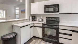A kitchen with white cabinets, a black microwave, a stove, and a trash can. - < 1 Mi to Ocean! OBX Condo w/ Decks, Pets Welcome (Hatteras)
