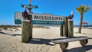 Ein hölzernes Schild mit der Aufschrift 'Mississippi Gulf Coast' und einem Willkommensschild, eine Holzbank und eine Palmenbaum.