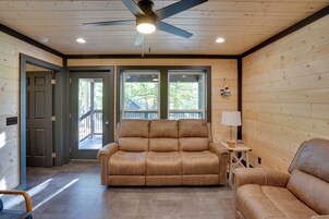 Una habitación con techo de madera, ventilador de techo, sofá, una silla, una pequeña mesa con lámpara y una puerta que da al exterior.