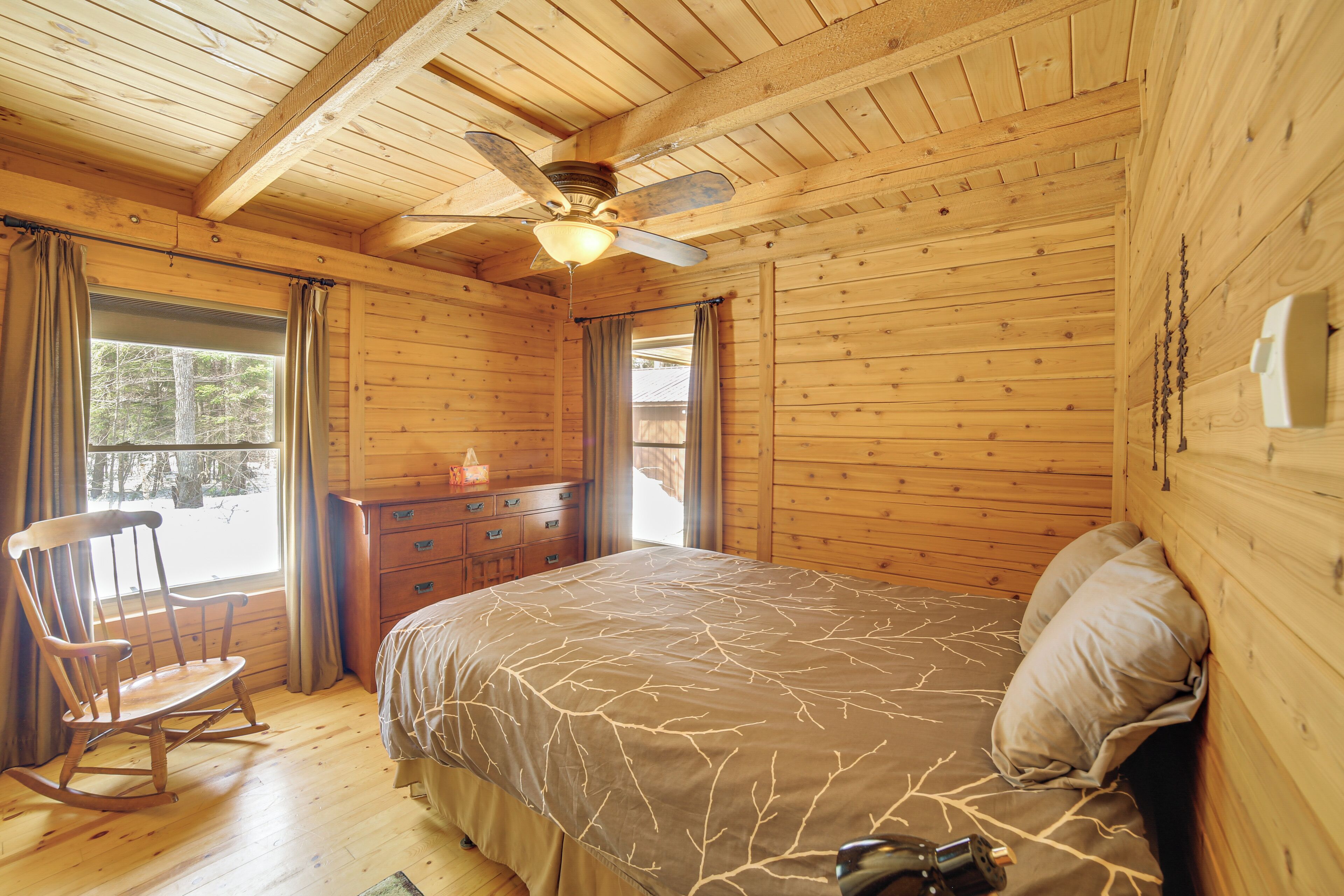 Une chambre dans une cabane en bois, avec un lit, un fauteuil à bascule, un ventilateur de plafond et une commode.