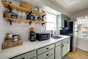 A kitchen with a microwave, coffee maker, and a refrigerator. There are wooden shelves with glassware and a basket. - Walkable Home in Dtwn Caldwell! 1 Mi to Campus (Caldwell)