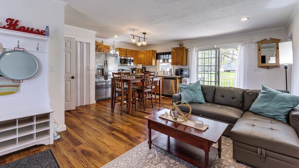A spacious living room with a sectional sofa, a dining area with wooden chairs, a kitchen with wooden cabinets, and a large window. - Dog-friendly Waneta Lake Home w/ Lake Access! (Hammondsport)