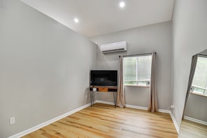 Une pièce avec un plancher en bois, un bureau avec une chaise, un appareil de climatisation et une fenêtre avec des rideaux.