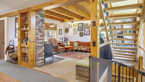 A spacious living area with a wooden staircase, a brick fireplace, and a bookshelf filled with books and decorative items. - Catskills Serenity! Cozy Vintage Cabin on 10 Acres (Grahamsville)
