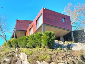 A modern two-story house with a red exterior, situated on a hillside with surrounding vegetation. - Casa Santa Luzia (Âncora)