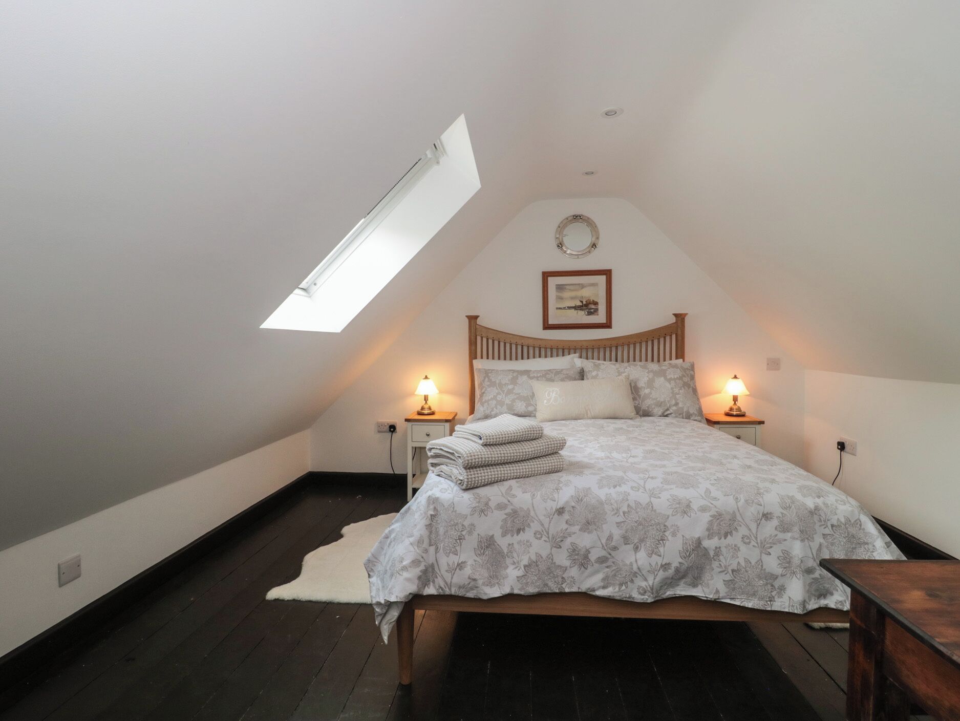 A bedroom with a skylight, a bed with floral bedding, two bedside tables with lamps, and a framed picture on the wall.