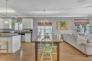 Eine moderne Küche mit weißen Schränken, ein Essbereich mit Holztisch und bunten Stühlen sowie ein Wohnzimmer mit Sofa und großem Fenster.