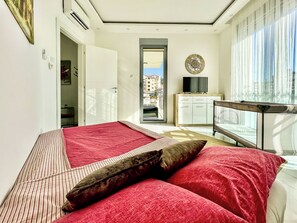 Ein Schlafzimmer mit Bett, Fernseher, einer Uhr und Blick auf Gebäude durch das Fenster.