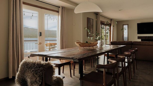 Uma área de jantar com mesa de madeira, cadeiras, um vaso branco com flores e vista para uma varanda com mesa e cadeiras do lado de fora.