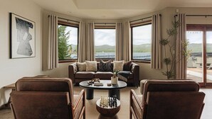 Sala de estar com sofá, cadeiras, mesa de centro e vista para um lago e montanhas através de amplas janelas.