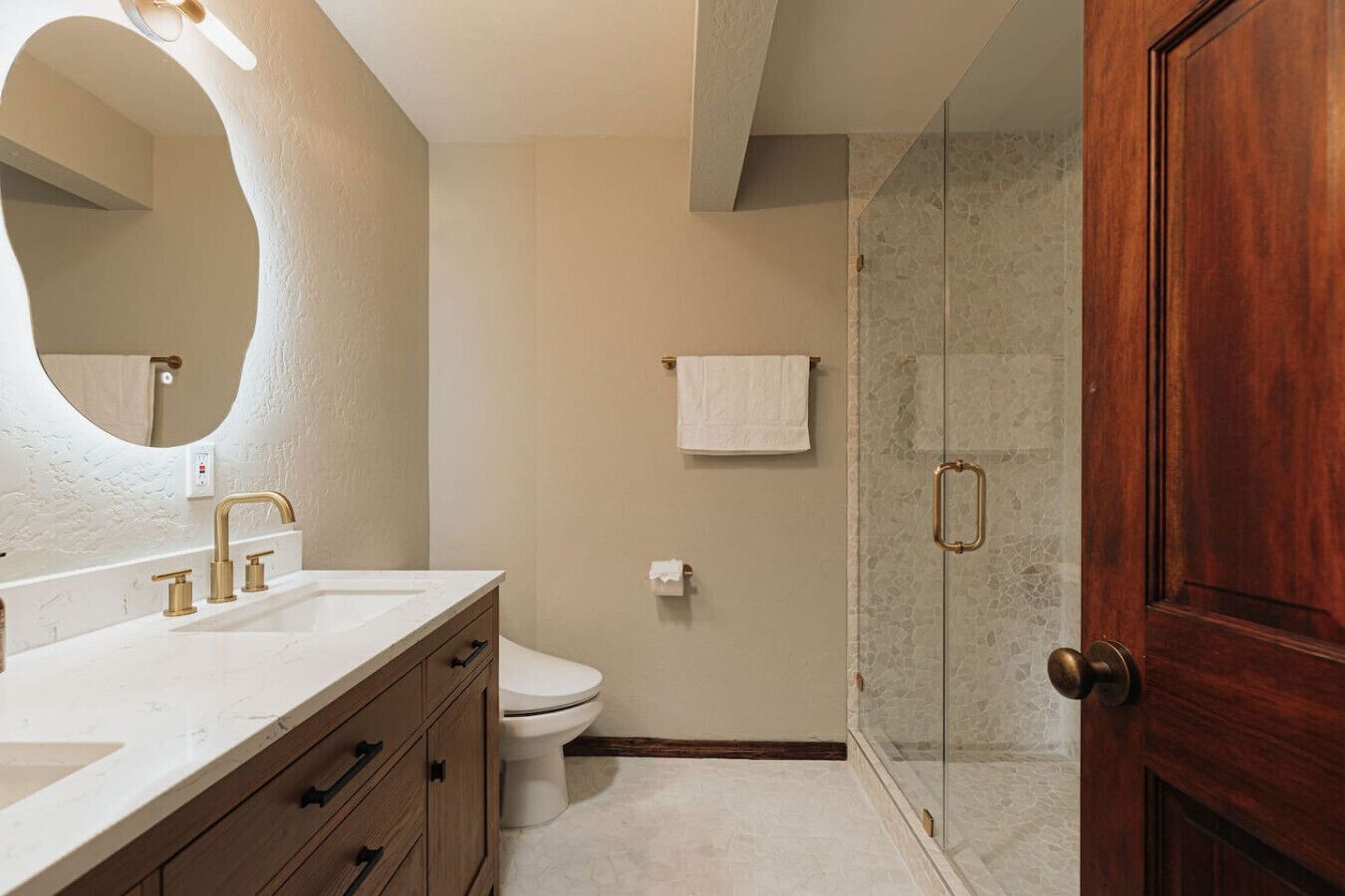 A bathroom with a white sink, gold faucets, a wooden vanity, a white toilet, a glass shower enclosure, and a wooden door.