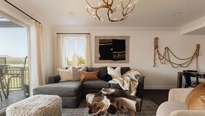 Een woonkamer met een hoekbank, een kroonluchter, een vloerkleed en uitzicht op de omgeving.