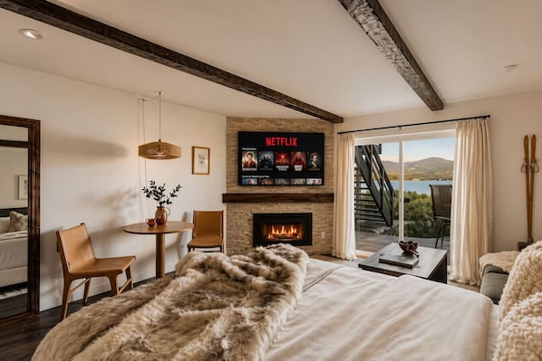 A bedroom with a fireplace, a TV mounted above it displaying Netflix, a bed with a fur throw, a dining area with chairs, and a view of the outdoors through a large window.