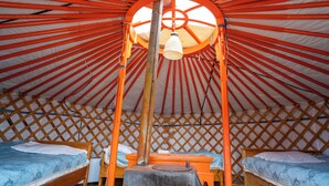 A yurt with a central wooden pole, two beds with blue bedding, and a hanging light fixture.