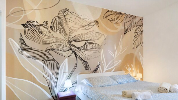 Um quarto com uma cama grande, uma mesinha de cabeceira, uma parede com um mural floral e um banquinho roxo pequeno.