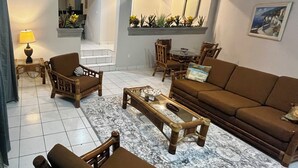 Ein Wohnzimmer mit Sofa, Sesseln, einem Couchtisch und einem Teppich. Eine Treppe führt zu einer oberen Ebene, eine Lampe und ein Pflanzenregal sind ebenfalls vorhanden.