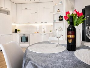 A modern kitchen with white cabinetry, a dining table with placemats, a wine bottle, and a vase with red flowers.