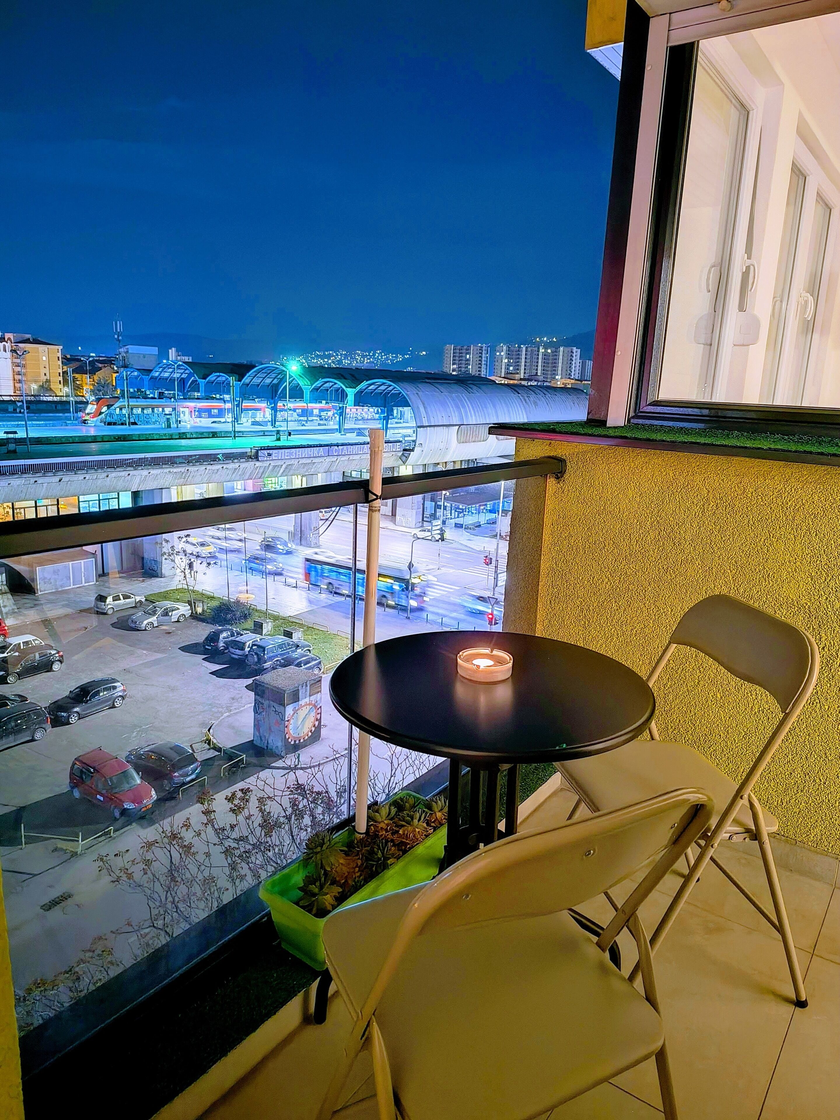 A balcony with a table and chairs, overlooking a parking area and a train station.
