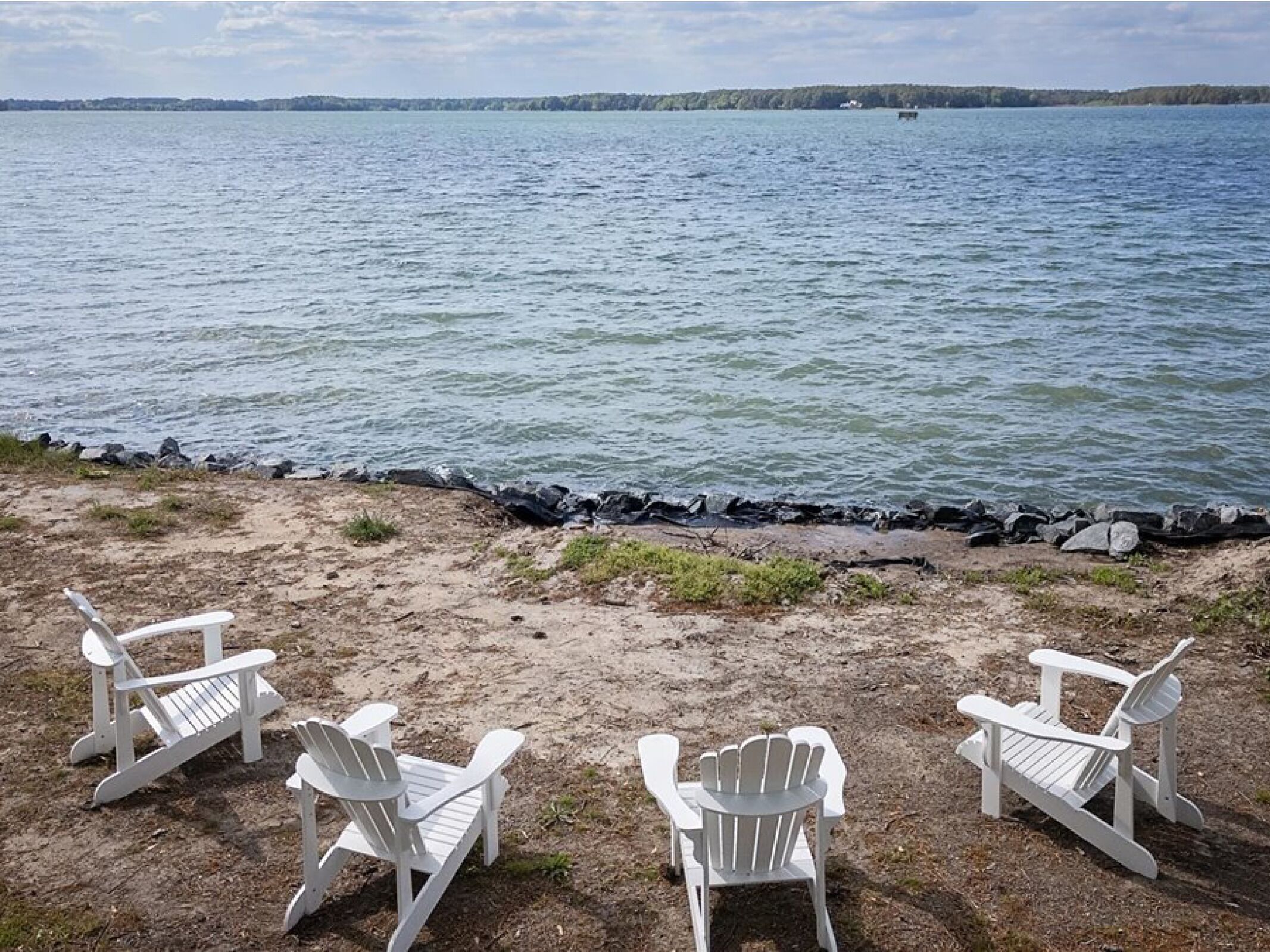 Trois chaises Adirondack blanches sont disposées face à un lac paisible, dont les rives sont rocheuses et qui est bordé d’arbres à l’horizon.