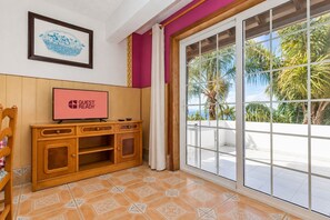 Una habitación con una televisión mostrando 'GUEST READY', un mueble de madera, un piso estampado y vistas a palmeras a través de una puerta de cristal.