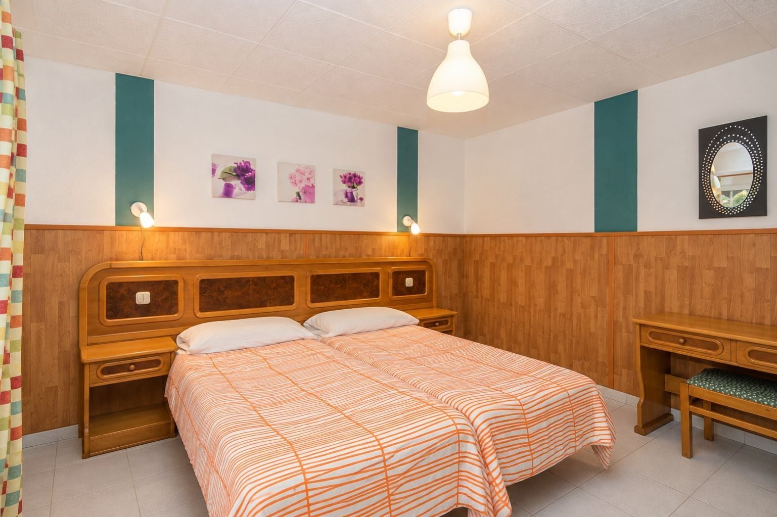 Una camera da letto con una testiera in legno, un letto con copripiumino a righe arancioni, un comodino, uno specchio e lampade a parete.