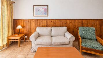 Ein Wohnzimmer mit einer holzverkleideten Wand, einem beigen Sofa, einem grün gemusterten Sessel, einem Holztisch und einer Lampe auf einem Beistelltisch.