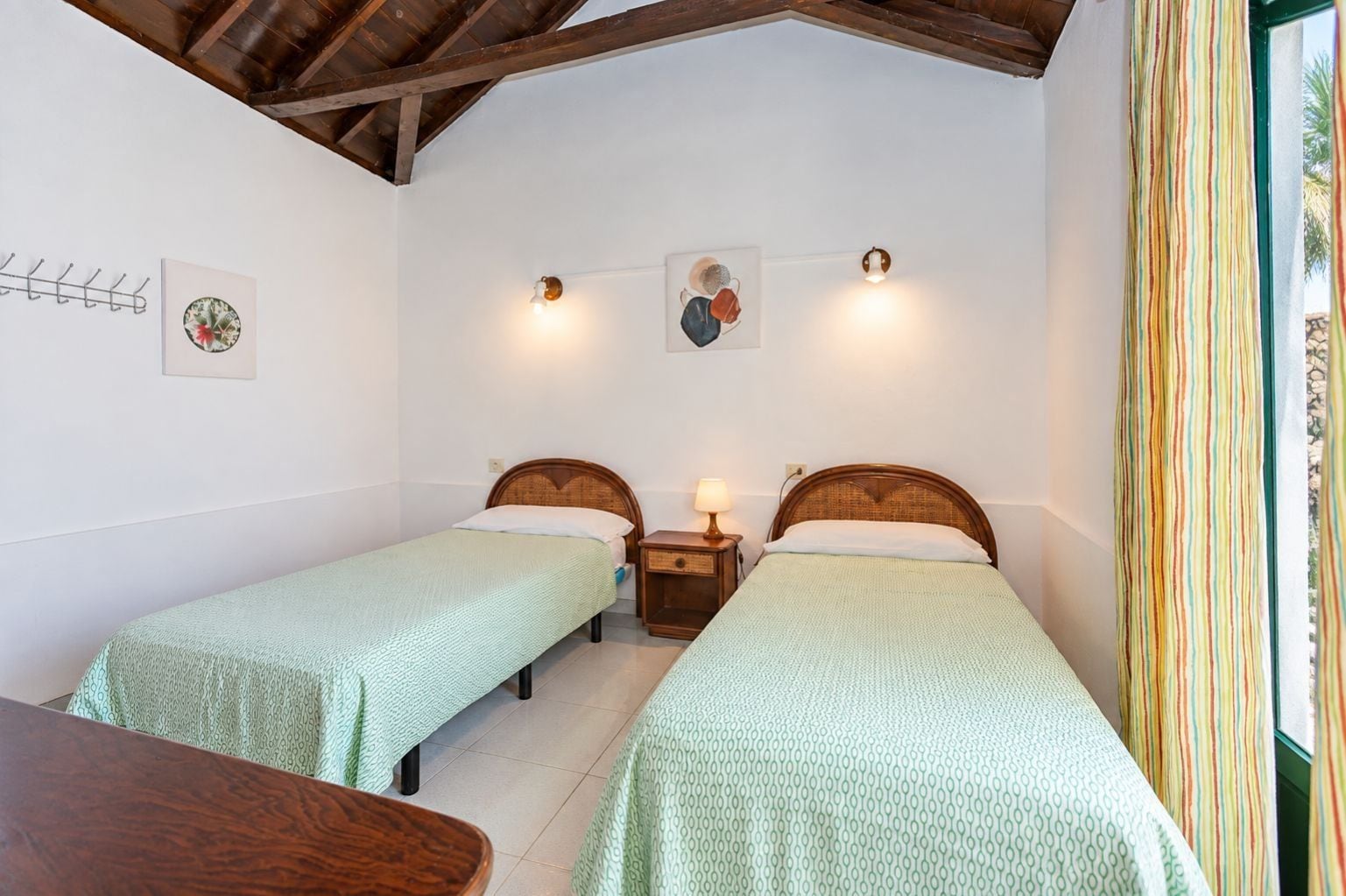 Una habitación con dos camas individuales, una mesa de madera, una lámpara de pared y un ventanal con cortinas.