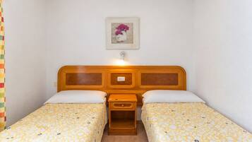 A small, single-room accommodation with a wooden headboard, two beds with matching bedding, a bedside table, and a wall-mounted light fixture.