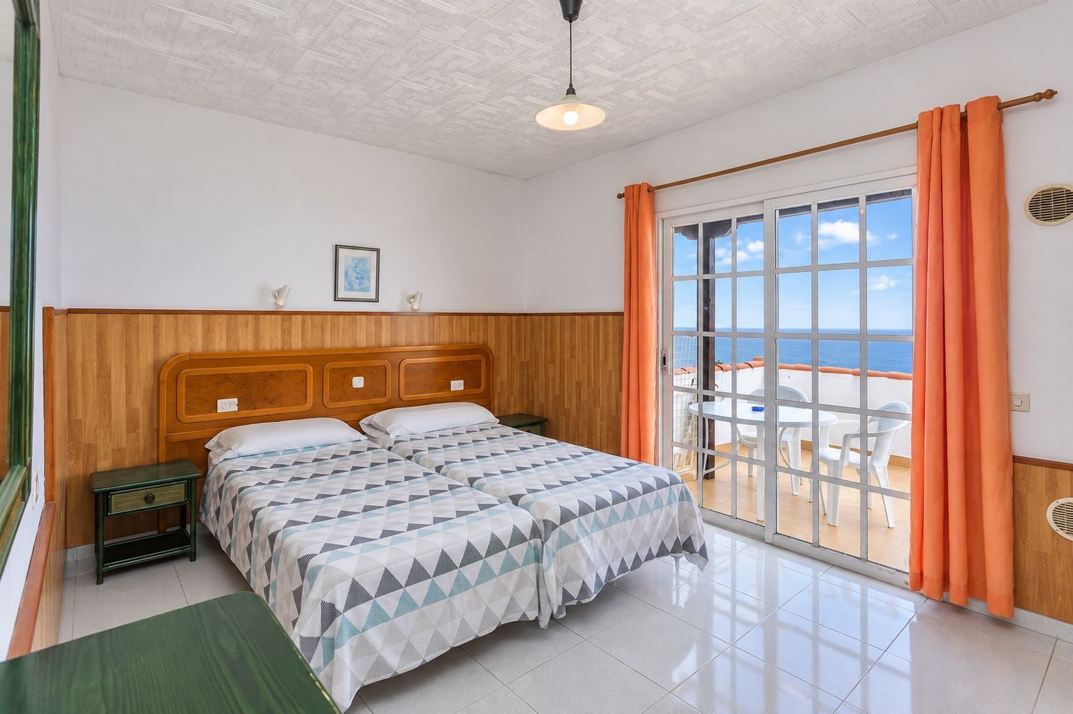Un dormitorio con cabecera de madera, cama con colcha de estampado geométrico, mesita de noche y vista al mar a través de una puerta corrediza de vidrio.