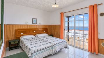 A bedroom with a wooden headboard, a bed with a geometric-patterned bedspread, a nightstand, and a view of the sea through a sliding glass door.