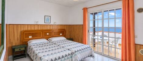 Un dormitorio con cabecera de madera, cama con colcha de estampado geométrico, mesita de noche y vista al mar a través de una puerta corrediza de vidrio.