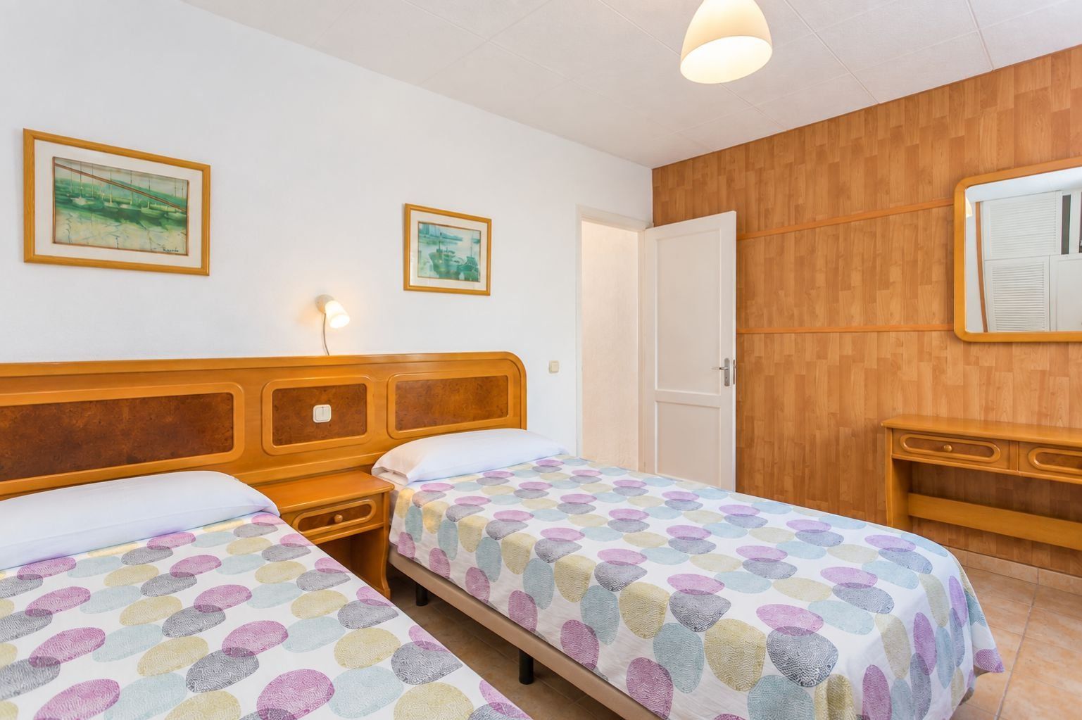 A hotel room with two beds, a wooden headboard, a bedside table, a nightstand, a wooden dresser, a mirror, and two framed pictures on the wall.
