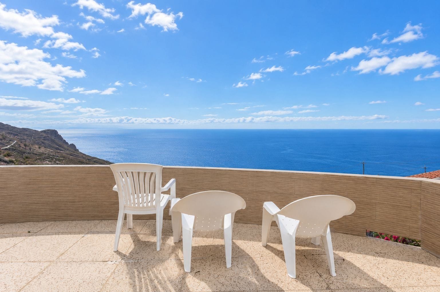 Una terrazza con due sedie bianche e un tavolo, affacciata su un paesaggio costiero sotto un cielo sereno.