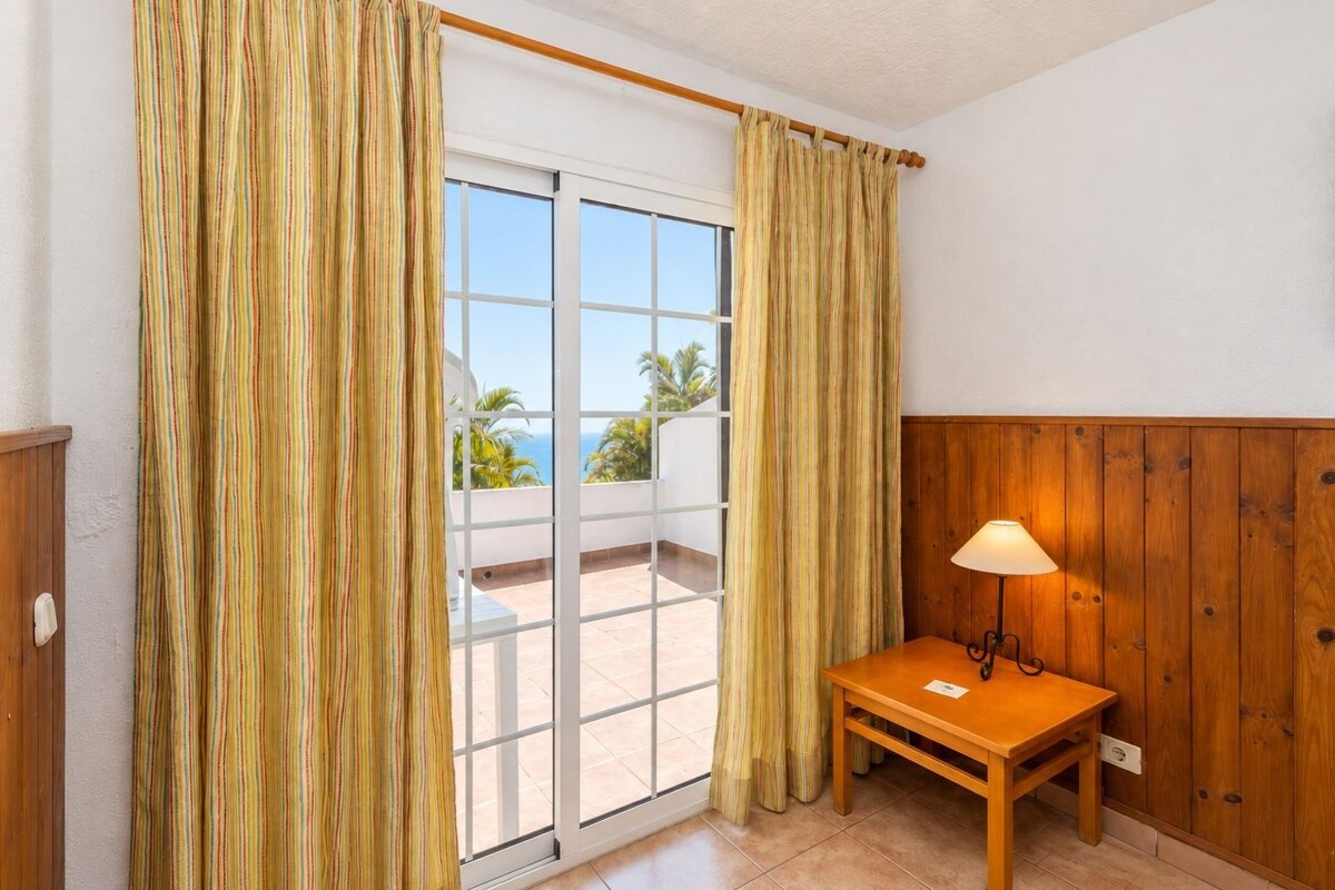 A room with a wooden table, a lamp, and a view of the ocean through a sliding glass door.