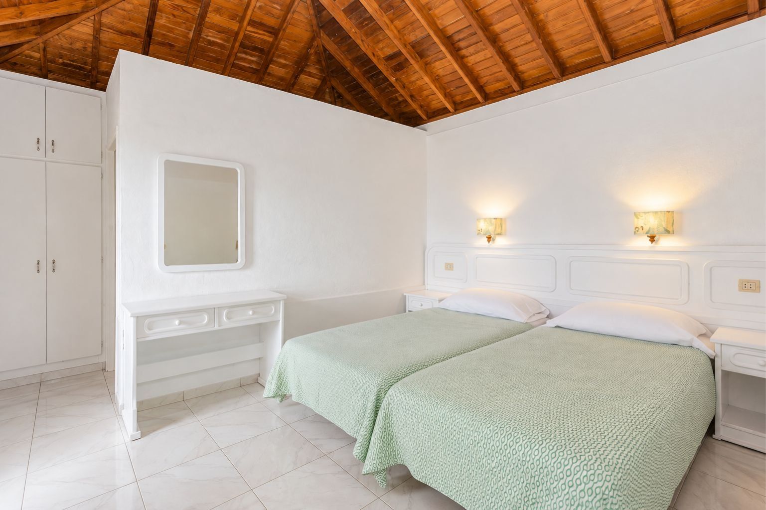A bedroom with a wooden ceiling, white walls, a bed with green bedding, a dresser, a mirror, and wall-mounted lights.