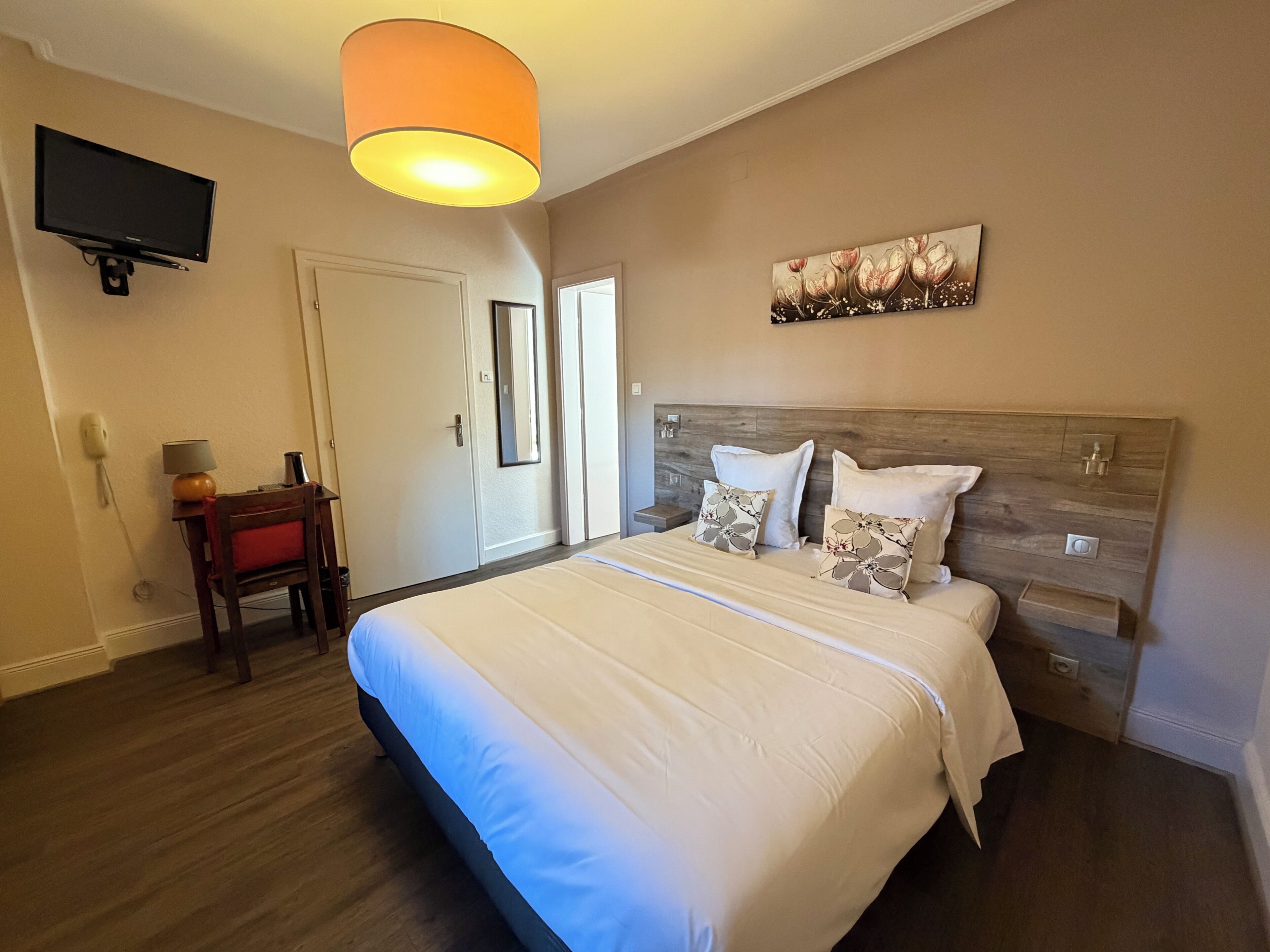 Quarto de hotel com cama, televisão presa na parede, mesa de cabeceira e porta.