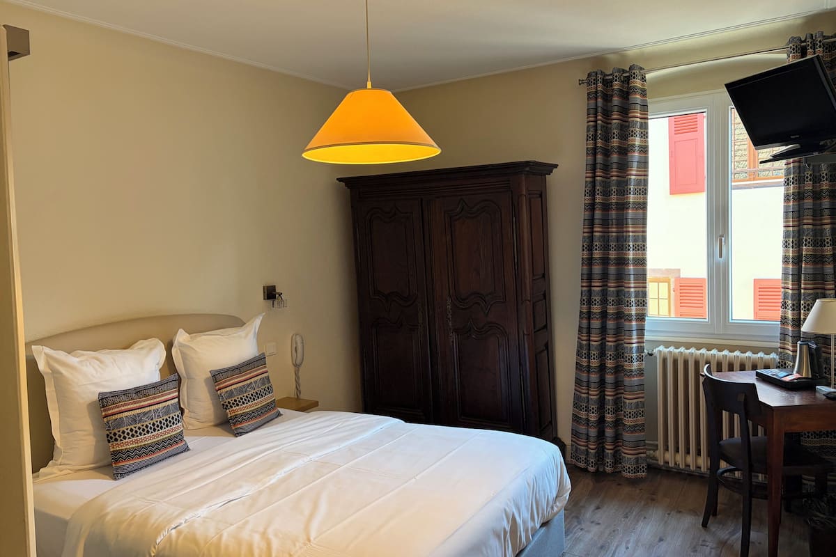 Un dormitorio con cama, armario, escritorio con silla, televisor y ventana con cortinas.