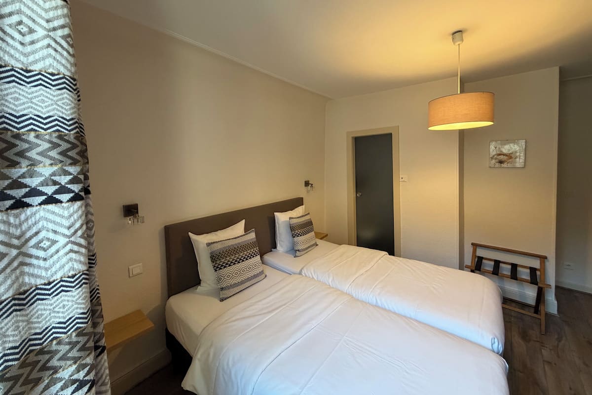 Una cama doble con ropa de cama blanca, una cortina estampada, una mesita de noche, una lámpara y una puerta en una habitación de hotel.