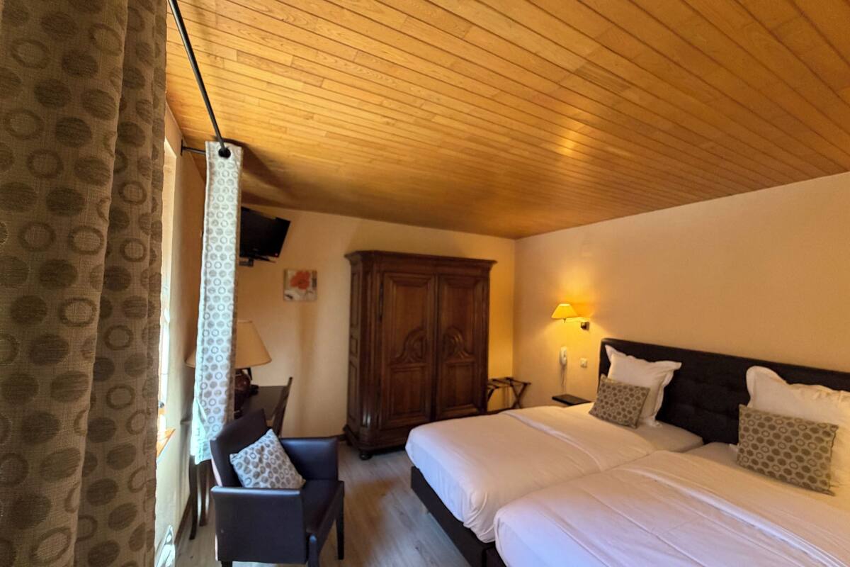 Un dormitorio con techo de madera, cama con ropa de cama blanca, armario de madera, escritorio con silla y una lámpara.