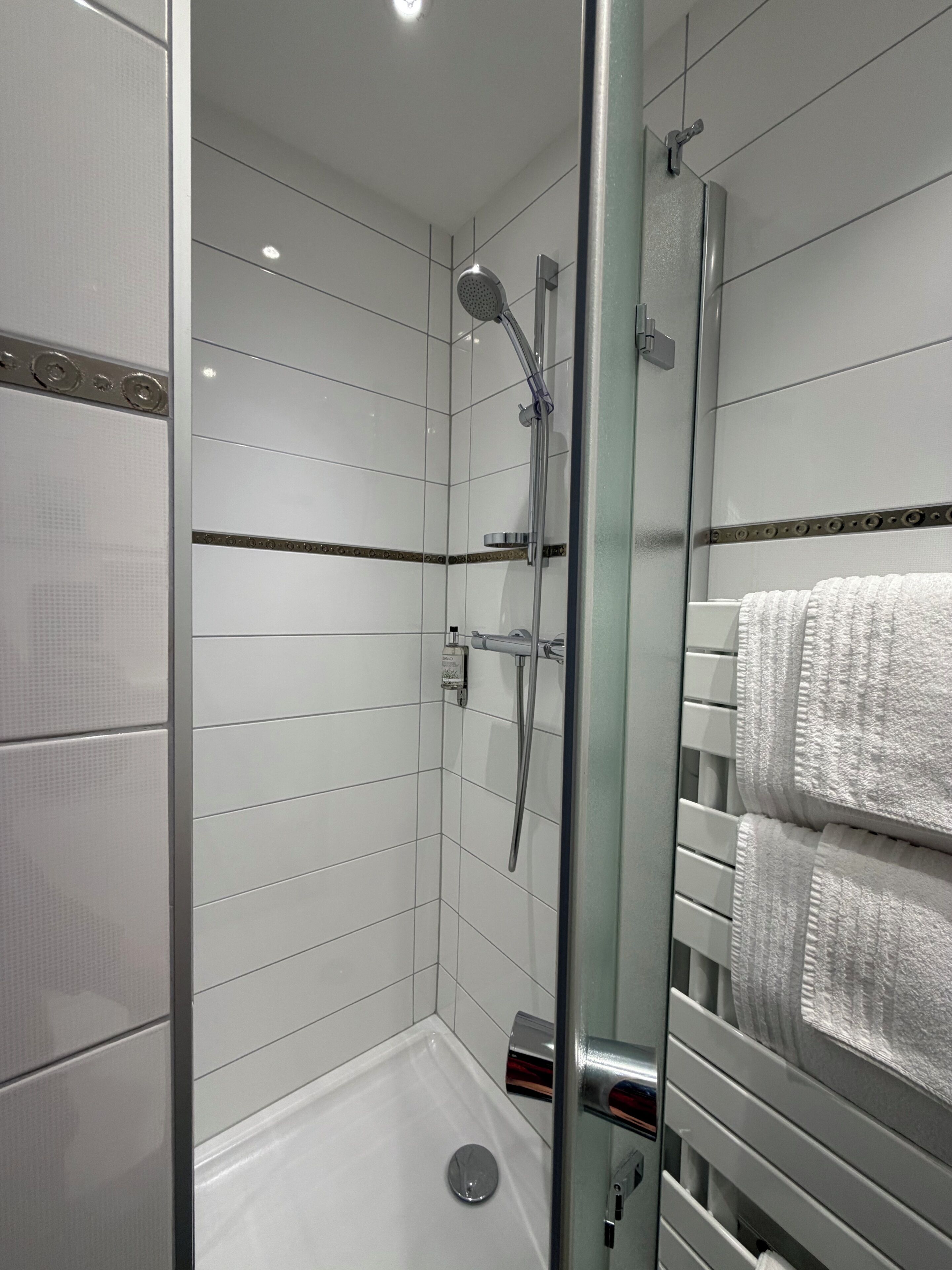 A modern shower with a glass door, white tiled walls, and a towel rack with folded towels.