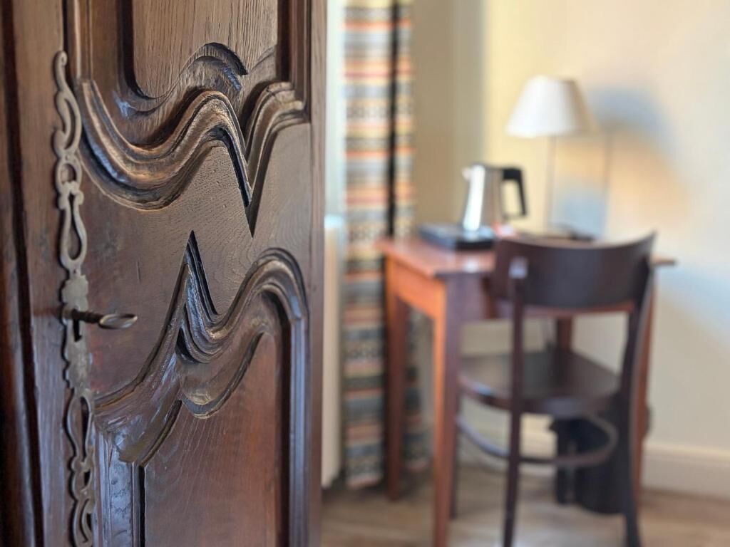 Uma porta de madeira com esculturas detalhadas, uma mesa com uma luminária e uma cadeira em um quarto com cortinas listradas.