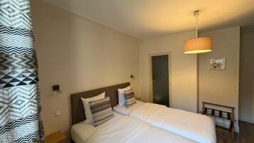 A hotel room with two beds, a bedside table, a lamp, a bench, and a door.