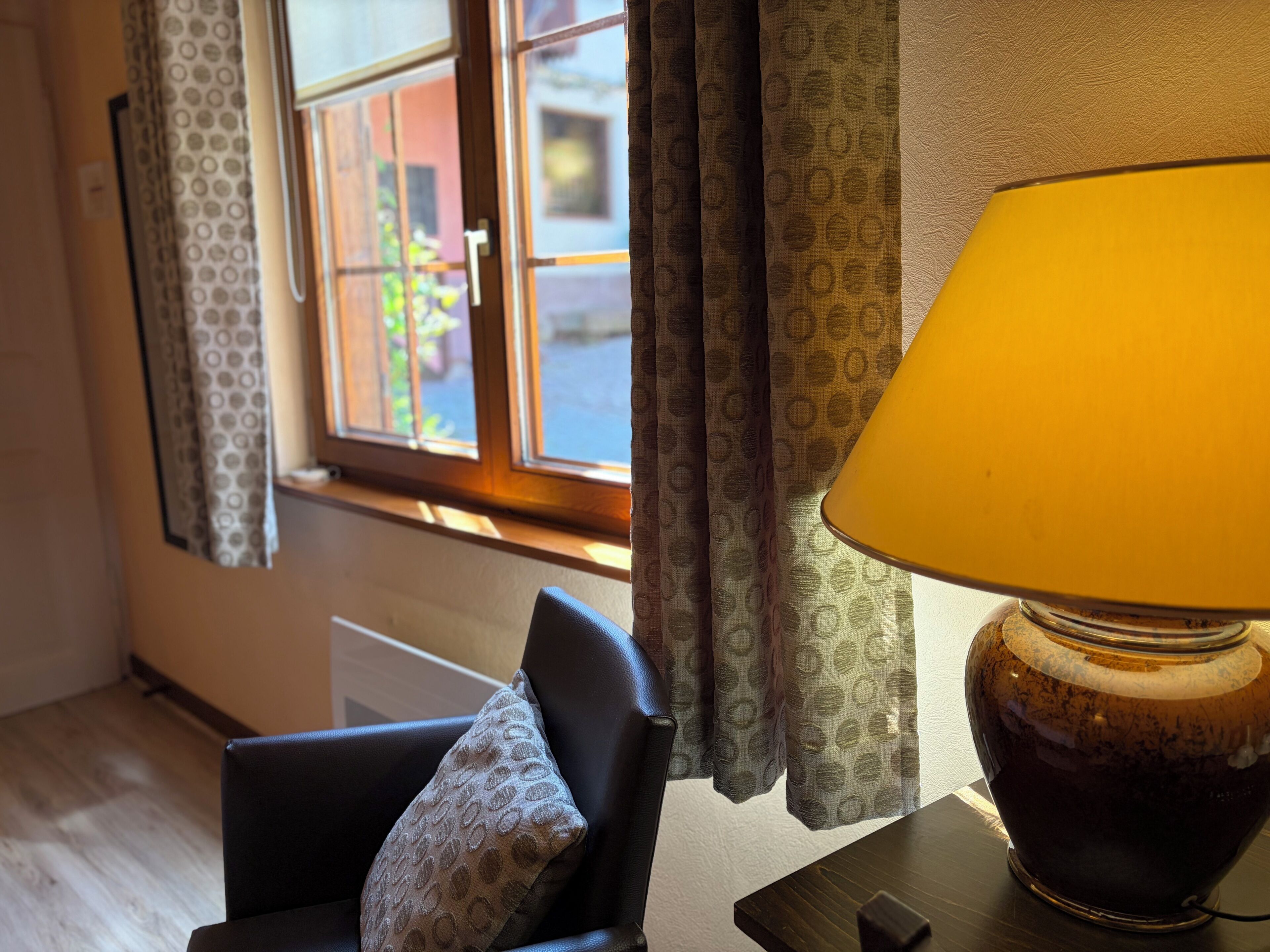 Una habitación con ventana, lámpara, silla y cortina estampada.