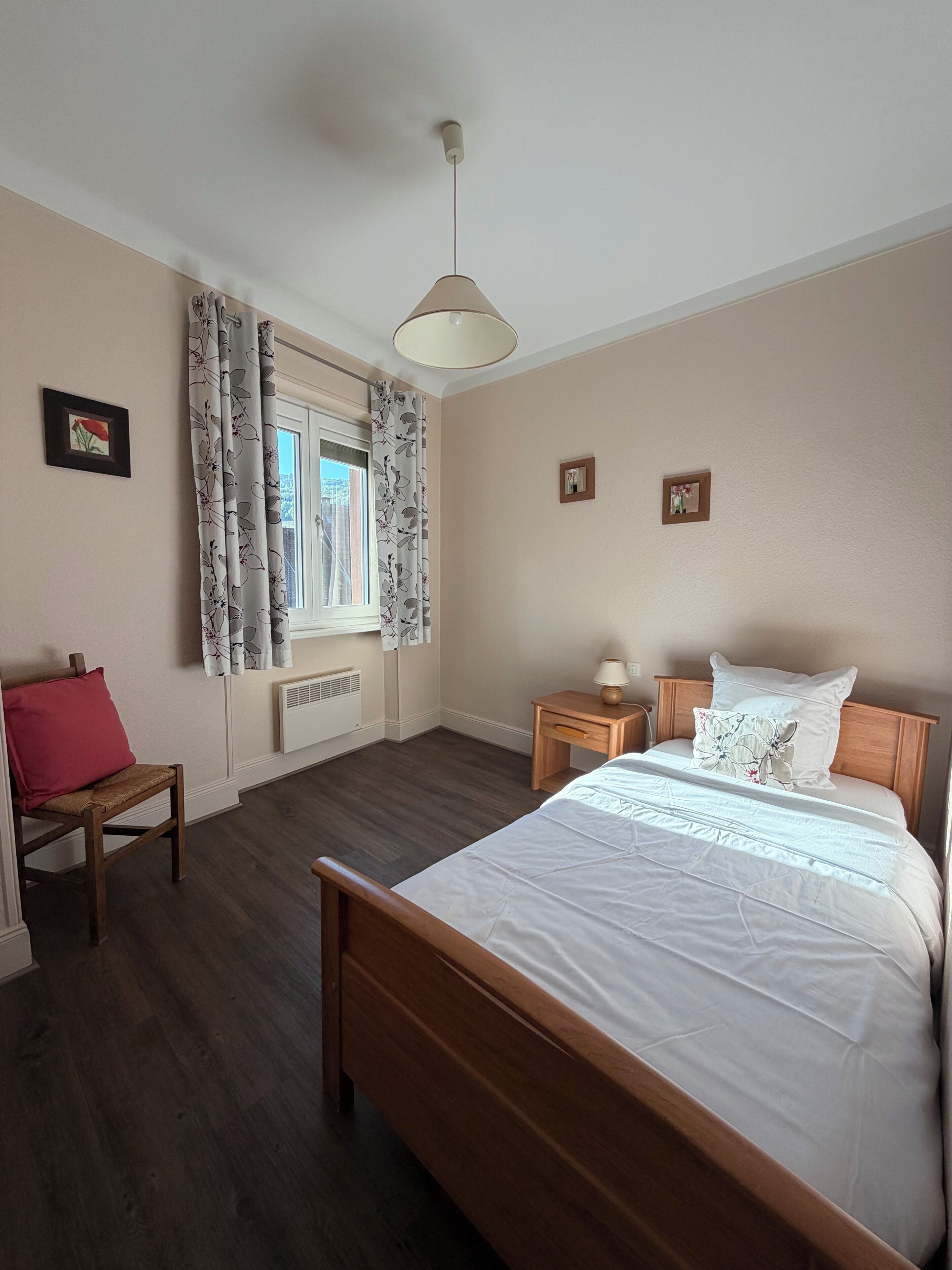 Un dormitorio con una cama de madera, una mesita de noche, una silla, una ventana con cortinas y una lámpara en el techo.