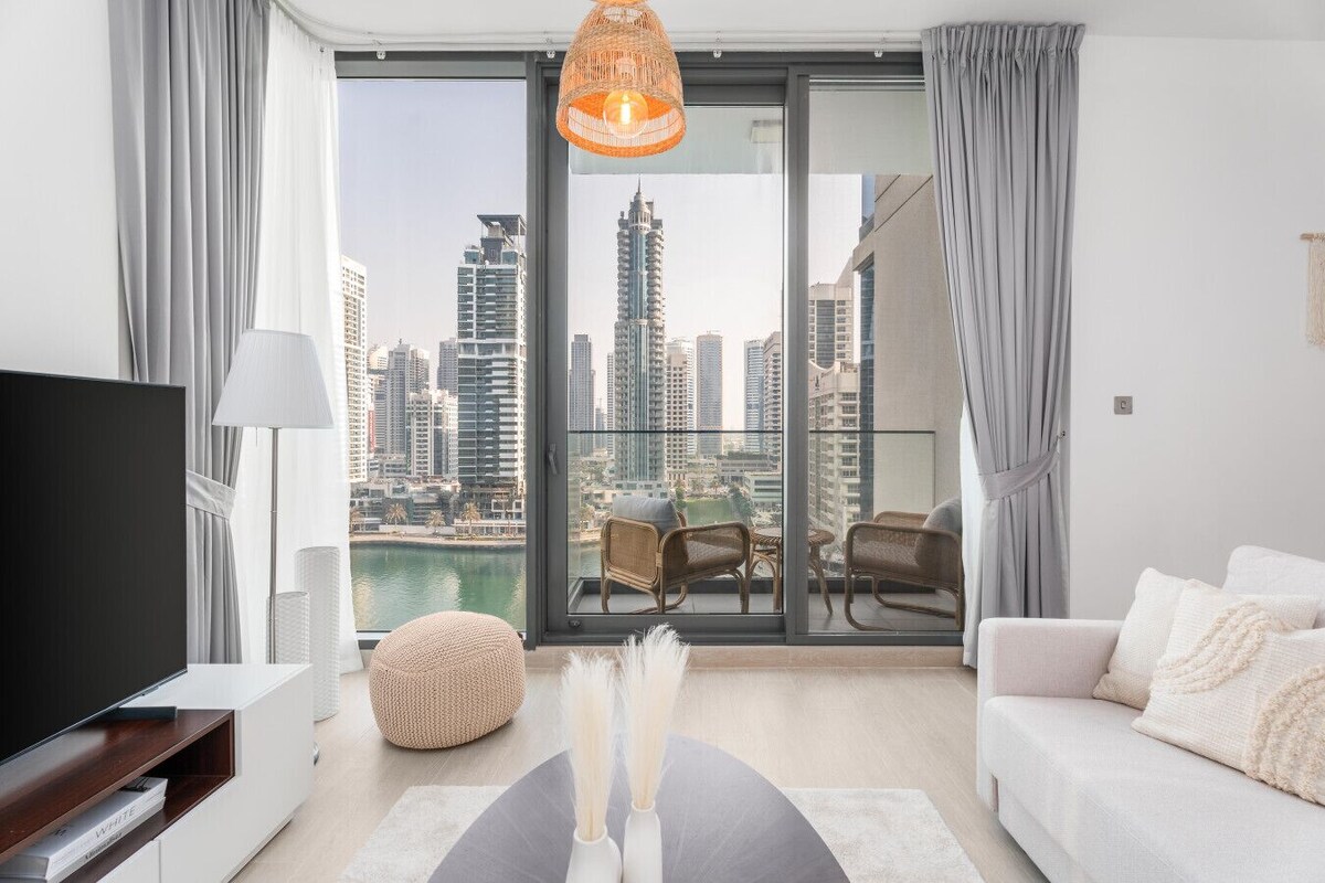 A modern living room with a large TV, comfortable seating, a view of a city skyline, and a balcony with a city view.