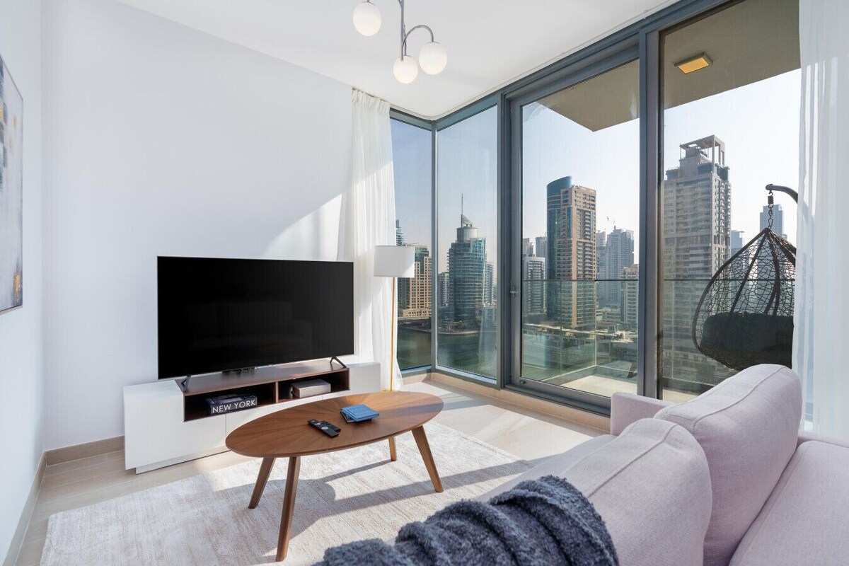 A modern living room with a large TV, a sofa, a coffee table, and a city view through floor-to-ceiling windows.