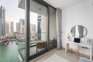 A modern room with a glass door leading to a balcony, a white desk, a chair, a round mirror, and a view of a cityscape and water. - Gala Guests - LIV Apartments Collection (Dubai)