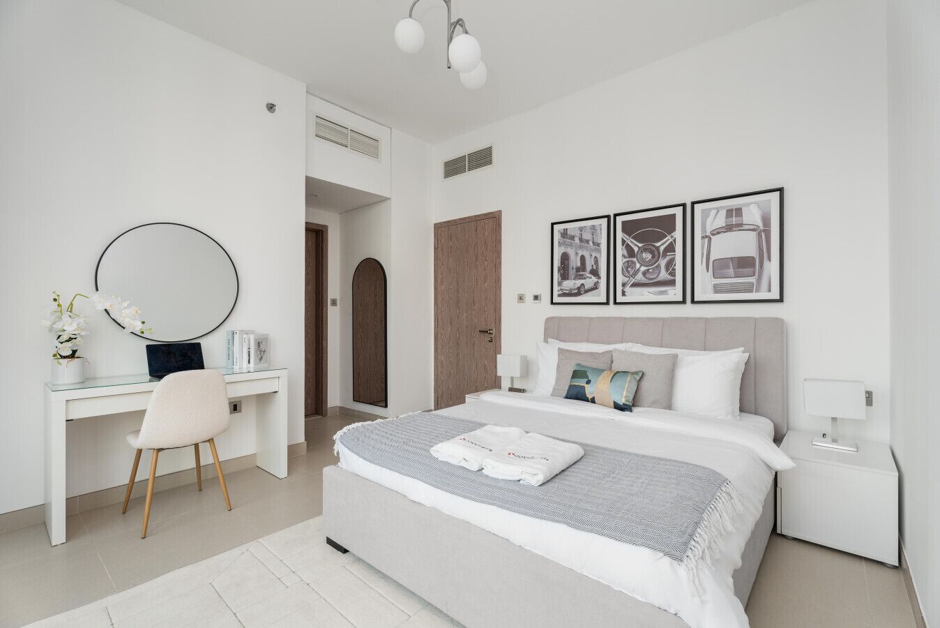 Une chambre moderne avec un grand lit, un bureau et une chaise. Des tableaux encadrés ornent le mur, et une porte est visible au fond.