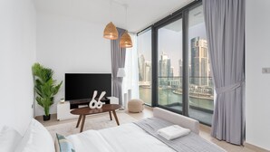 A modern bedroom with a large bed, a flat-screen TV, a potted plant, and a city view through floor-to-ceiling windows. - Gala Guests - LIV Apartments Collection (Dubai)